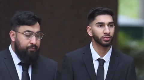 Brothers Muhammad Amaad (left) and Mohammed Fahir Amaaz who have black hair and beards and are wearing black suits with white shirts and ties. Muhammad Amaad wears glasses. The pair are not smiling.
