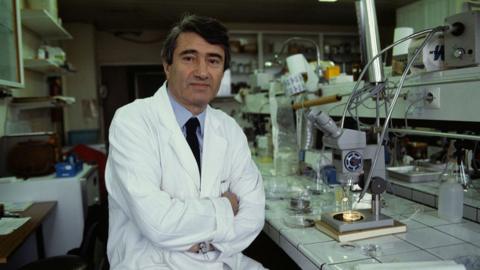 Dr Etienne-Emile Baulieu sits in a white lab coat with his arms crossed over his waist inside a lab, with various equipment around him and in the background
