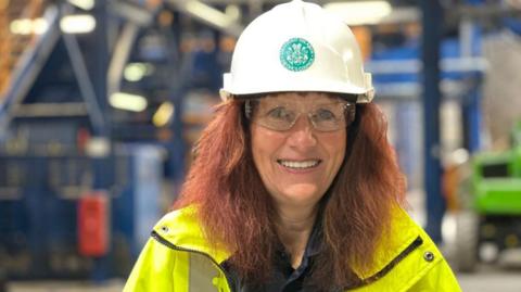 A smiling woman with long ginger hair wearing a yellow reflective jacket, a white helmet and protective glasses in a room with machinery in the background