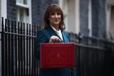 Rachel Reeves stands wearing a blue suit and white shirt, holding the red budget box.