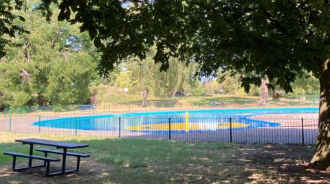 A blue and yellow splash pool in a park. It is surrounded by a grassed area with large green trees and a green metal fence.