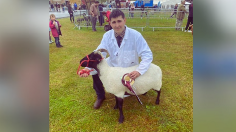 Will Sully at an agricultural show, standing with Knobby, who has just won a dark red and gold rosette at the show.