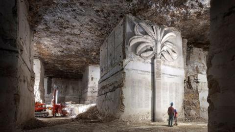 An artist's interpretation or illustration of the Memo Portland project. It shows a carving of a large tree on a white block of stone inside of a disused mine, with words inscribed on the trunk of the tree. Beneath it two people are looking up at the top of the tree as it towers over them. A bulldozer can be seen in the background of the caverns. 