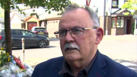 Maolíosa McHugh has short grey hair thinning on the top and glasses. He is wearing a navy blazer and brown shirt