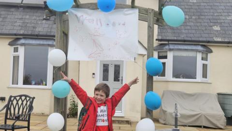 Riley wearing a red jacket and a red t-shirt which reads 'A Bear Named Buttony'. He is standing centre with his arms in the air in front of an archway decorated with blue and white balloons and a sign which says 'Well done Riley from Lucy'. A house is in the background.