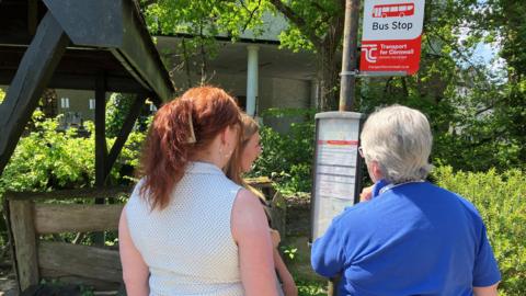 Three women with their backs to the camera are looking at the bus stop at Trago Mills in Liskeard looking at the bus timetable 