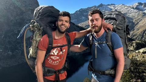 Two men stand with their arms on each others' shoulders near a river in a mountainous area - both have packs on their backs and wear T-shirts and are smiling, it is a sunny day 