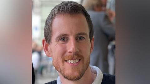 PC Miller with short brown hair and beard smiling at the camera. He is wearing a blue sweater with a white neck