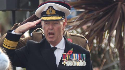 Jonathan Carley dressed as an admiral gives a salute. He wears a white hat, navy jacket, white shirt and tie. On his chest are a series of medals hung by colourful ribbons. 