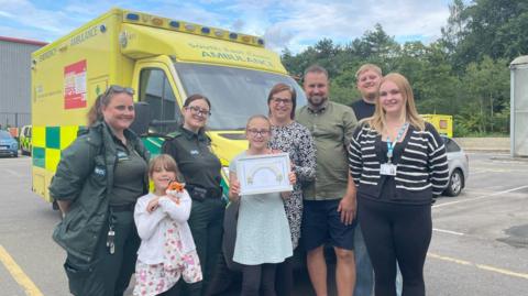 Alice holds her certificate of bravery, while standing next to her sister. She is surrounded by her mum, her dad, a chief executive of a hospital trust, another man and two paramedics. An ambulance is in the background.