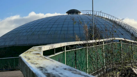 The Oasis Leisure Centre exterior. There is a large dome with a ladder running to the top. There is a pavement with a green railing running across to the right.