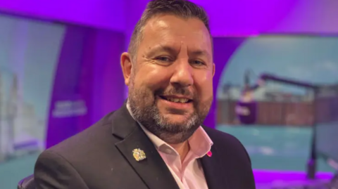Anthony Burns sits in a radio studio, a blue microphone and control desk are visible in the background. He smiles at the camera. He has short dark hair and a stubbly beard. He wears a dark suit jacket with a pink, open-collar shirt underneath.