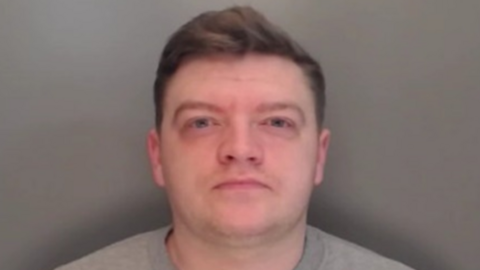 Mugshot of Jordan Stoker. He is clean shaven with a wide face and eyes set far apart from each other. He has short dark hair and is wearing a grey sweater