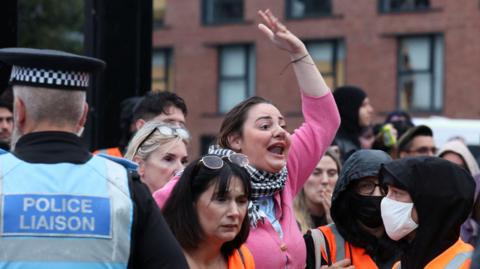 One protester, wearing pink, holds her arm up in the air and shouts as a police liaison officer stands to the left and crowds surround the woman