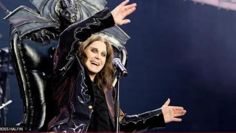 A long-haired man sits in a throne with a black bat design behind him. He is wearing a purple-ish velvety looking coat, is waving his arms and smiling. He has thick black eyeliner.