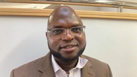 Dr Salako with a short beard and glasses wearing a brown jacket