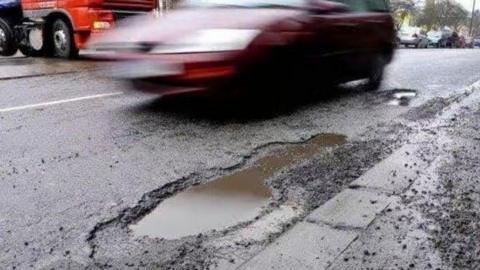 A red car which is blurry is driving past a large pothole on a road. The pothole is filled with water.