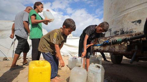 Two Palestinian boys fill up water cans, as a woman drinks water in the background