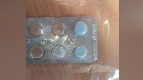 Blister pack with two blue pills inside. The pack is on top of a clear plastic bag on a wooden table. Six blisters can be seen but only two have pills in them.
