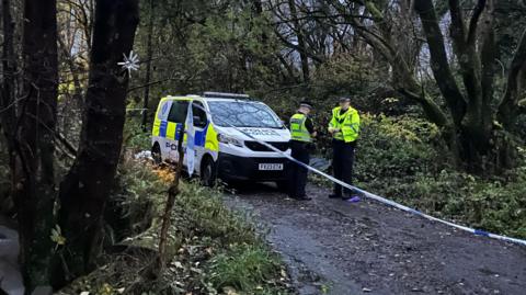 Two police officers wearing hi-vis clothing stand behind blue and white police tape in a wooded area. A marked police van is parked to their left.