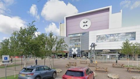 The front of the Xscape leisure complex in Castleford, pictured from the view of the front of the car park. The entrance has several tiers. A series of picnic tables and trees stand outside. 