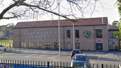 An image take over a short metal fence of a large brown brick building with the words GREENFIELD SCHOOL on the side. Between the fence and building is a large car park with three vehicles parked in it.
