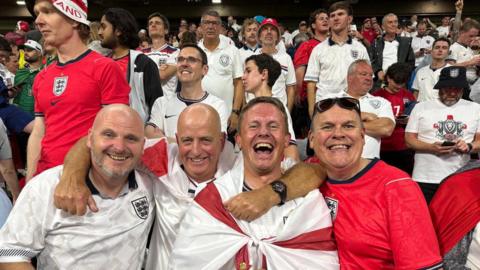 Jersey football fans Paul Starkey, Dave Houze, Paul Wilson, and Tony Rogers at the Azteca Stadium in Mexico after England's World Cup win. They are smiling and are wearing red and white England shirts. Dave Houze has a red and white England flag round his shoulders. Other England fans can be seen in the stadium behind them.