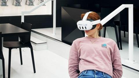 A woman with ginger hair and wearing a pink jumper tries out the new VR headset while sitting on a chair.