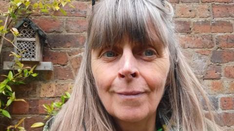 A woman with long brown hair stood in front of a brick wall with a plant growing up it