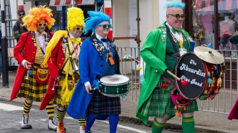 Four men in brightly coloured coats and wigs wearing kilts and holding drums.