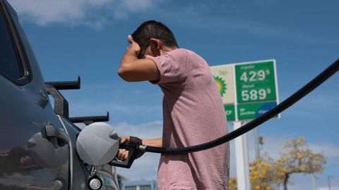 A man palms his forehead in frustration while filling his truck up with gasoline. 