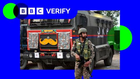 An Indian soldier stands guard. He is armed with a rifle and is wearing army fatigues. In the background is a large truck.