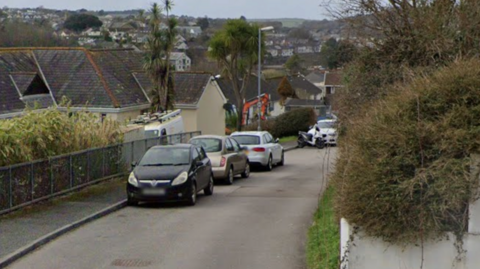 A Google Street View of Polruan Road in Truro. It is a single lane road surrounded by houses, and with cars parked on the pavement. In the background is a view of Truro.