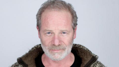 Peter Mullan, with greying beard and moustache, stares intently at the camera, a slight smile on his face. He wears a black t-shirt and a borg fleece jacket in shades of brown and cream