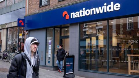 Man in a hoody walks past a Nationwide branch with another man walking out of the door.