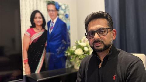 A man, Miten Patel, in a dark jumper, looking into the camera. In the background is a large image of two people in formal attire and a bouquet of white flowers. 