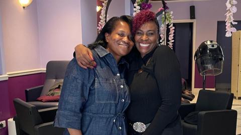 Wilma Burke smiles brightly alongside her client Katherine Kidd at Pride Hair and Beauty Salon. Wilma wears a long black fitted shirt with a silver-buckled belt, black trousers and has burgundy hair. Katherine, in a denim shirt tied at the waist, has freshly styled shoulder-length black hair. Both women look directly at the camera, smiling.
