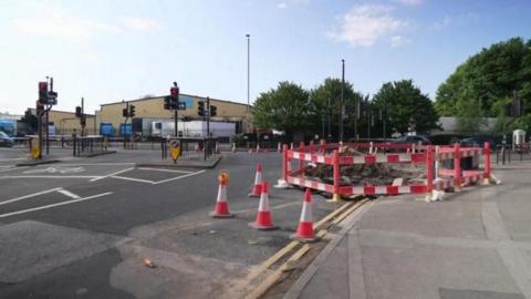 The junction of Princes Avenue and Spring Bank West in Hull. The road, which features multiple sets of traffic lights, is covered with orange and silver cones. A red and white barrier surrounds a large hole and a large pile of rubble. An industrial building stands in the distance.