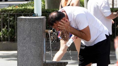 A man splashes his face with water at a drinking fountian in Spain during the heatwave. He is wearing a white T-shirt 