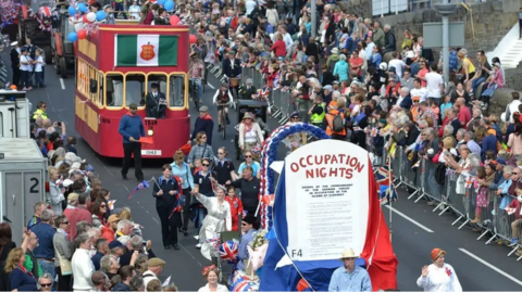 A view of the 70th Liberation Day cavalcade when floats were last included. One float is designed to look like an old red Guernsey tram and another is red white and blue and says Occupation Nights on it 