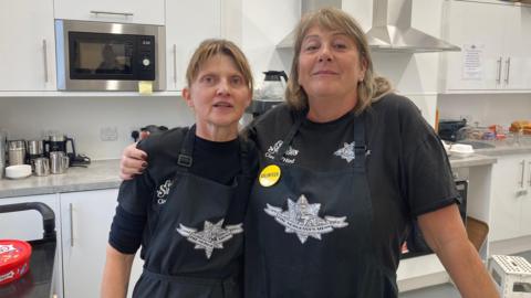 Two women wearing blue aprons, stood inside a cafe - behind them is the kitchen.