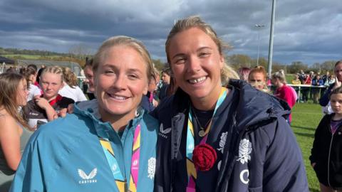 Red Roses Alex Matthews and Zoe Aldcroft in front of a pitch filled with young female rugby players.