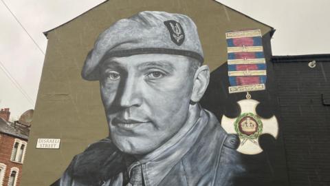 A mural of Blair 'Paddy' Mayne. He is painted in black and white and is wearing a beret with an SAS badge. On the right hand side of the wall is a Distinguished Service Order medal, a white cross with a gold crown in the middle suspended on a ribbon which has a pink stripe down the middle and blue stripes on each side.