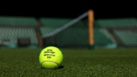 A bright yellow tennis ball sits on the grass on one of the courts at Wimbledon. Behind it is blurred but you can make out the net and the seats