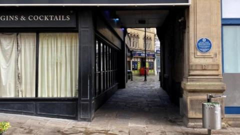 A short alleyway, about 10 metres long. The floor is paved with stone and the area appears clean. A betting shop inside an old stone building can be seen at the other end of the passageway.