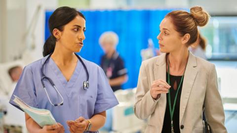 Stock image showing a female NHS consultant, dressed in scrubs, with a stethoscope around her neck and a folder in hand,  talking to a female administrator, wearing a tan jacket and with a green lanyard round her neck    