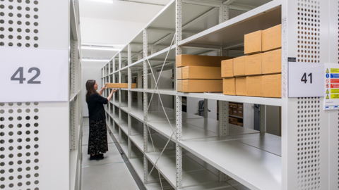 A woman holding a box in a room that has lots of metal shelves in it. There are numbered brown boxes on one of the shelves.