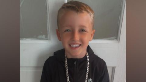 A six-year-old boy with light blonde hair, wearing a black coat with a large golden chain around his neck, grins at the camera while standing in front of a white wooden door. 