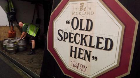 A large Old Speckled Hen beer sign on the side of a lorry, with three beer kegs, and a man bending down, wearing a high-vis jacket. 