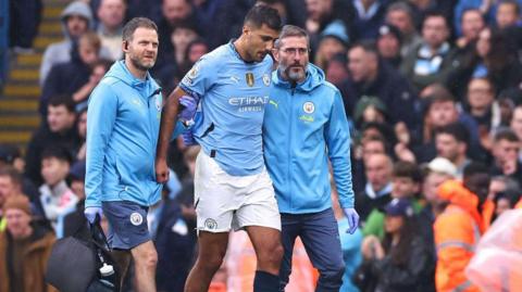 Manchester City midfielder Rodri being helped off the pitch by physios